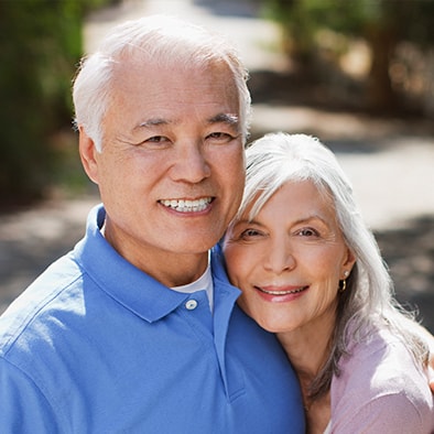 An older couple with dental implants smiling as the woman rests her head on her husband's shoulder