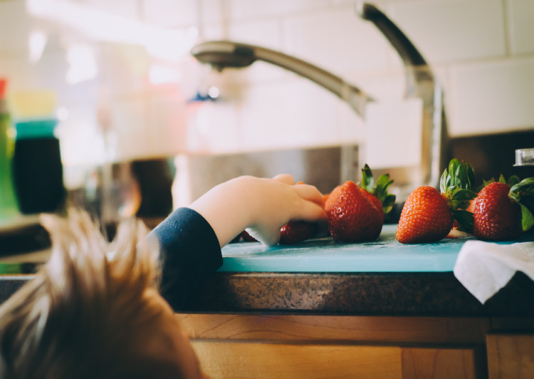 A blonde boy reaches for freshly washed strawberries on the kitchen counter because his parents teach him healthy eating