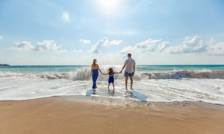 Mother, father, and daughter hold hands while standing in the shallow waves at the beach during summer vacation