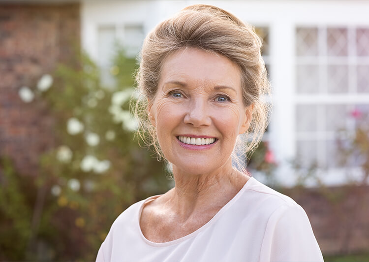 An older woman with dentures smiling in front of her house's rose bush.