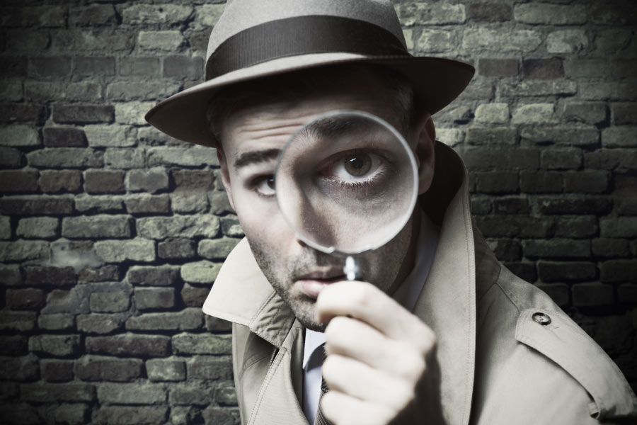 man in a fedora and overcoat holds a magnifying glass to his eye
