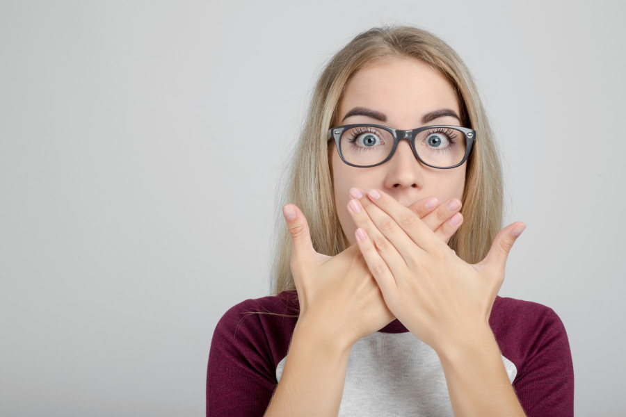 blonde woman wearing glasses covers her mouth with her hands to hide gum disease